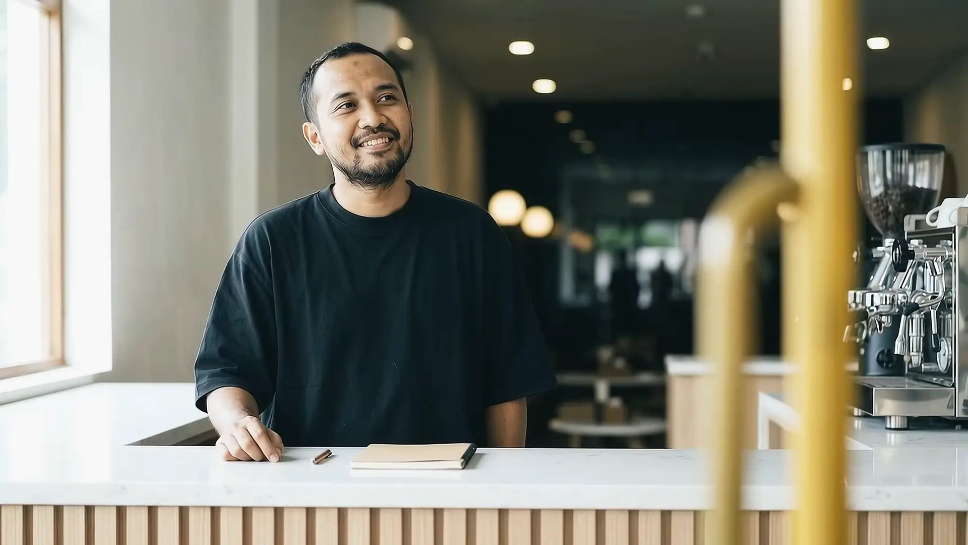 Pemilik kedai kopi tersenyum di balik meja kasir dengan buku catatan di depannya; lalu ia melayani pesanan di kasir Qasir dan menunjukkan aplikasi Qasir di HP-nya.